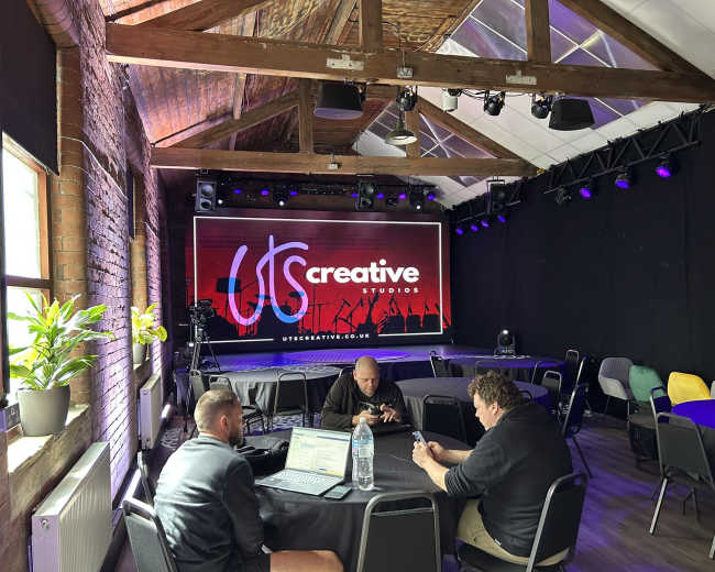A spacious studio with exposed brick walls and wooden beams features a large screen displaying the words "UTS Creative Studios," with several people seated at tables working on laptops.
