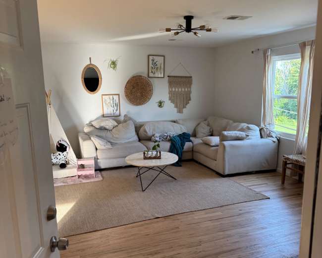 A bright living room features a light-colored sectional sofa, a round coffee table, and decorative wall hangings, seen through an open door.