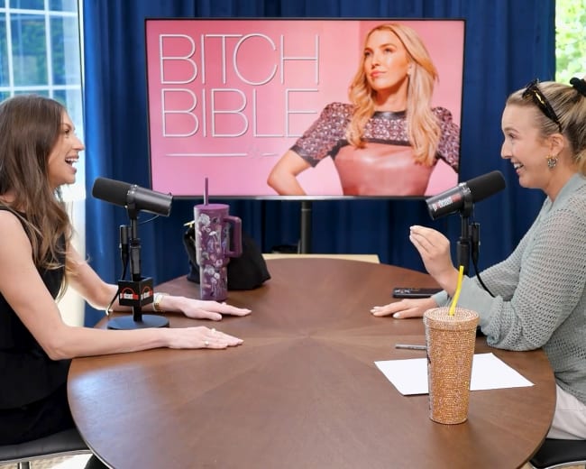 Two women are seated at a round table, engaging in conversation with microphones in front of them, while a large screen in the background displays the title "Bitch Bible" alongside an image of a woman.