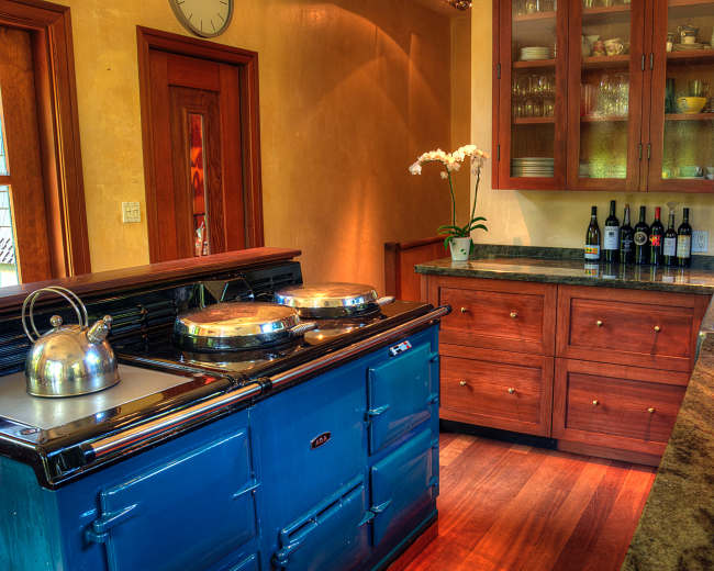 The image shows a kitchen with a blue stove, wooden cabinets, and a granite countertop, featuring a vase with an orchid and several bottles of wine on a shelf.