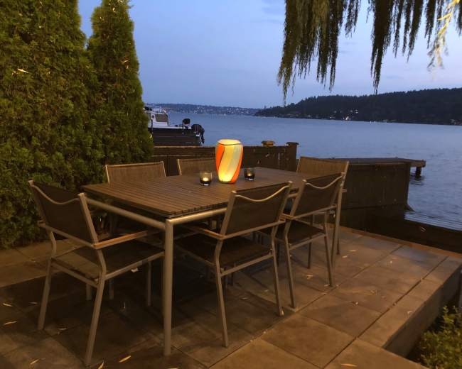 A wooden dining table with six chairs is set near the water's edge, illuminated by a lantern and overlooking a lake surrounded by trees and hills.