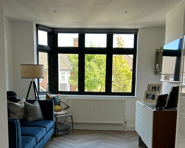 The image shows a modern living space featuring a blue couch, a lamp, a large window with a view of trees, and herringbone-patterned flooring.