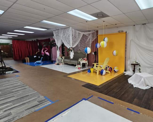 The image shows a studio space decorated with various backdrops, including a bright yellow wall, draped white fabric, beach balls, and props such as a ladder and chairs.