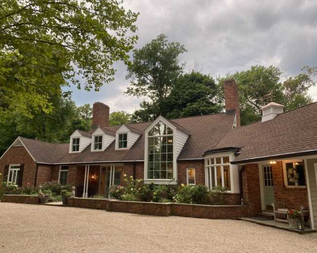 A large brick house with multiple gables and a central window is surrounded by trees and a gravel driveway.