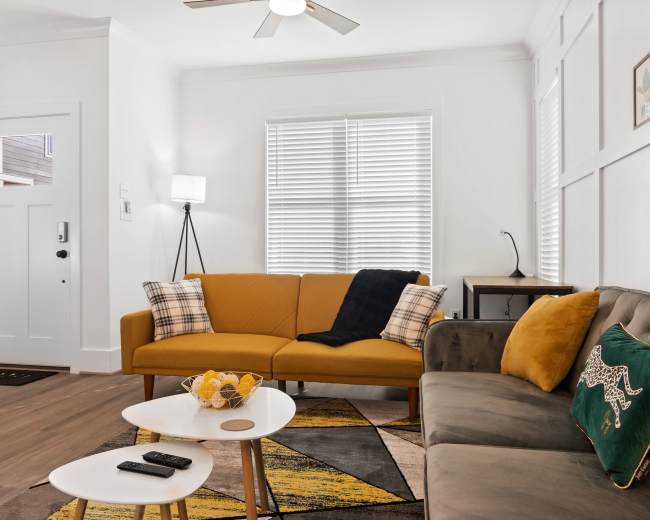 A modern living room featuring a yellow couch, a gray sofa, a coffee table, and a lamp, with large windows providing natural light.