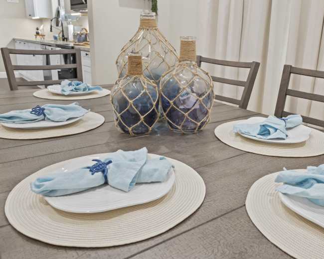 A dining table is set with white plates and light blue napkins arranged neatly on round placemats, featuring decorative glass vases in the center.
