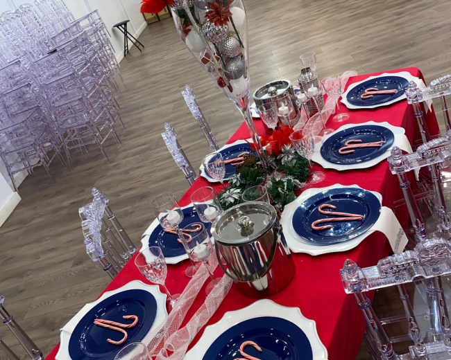 A decorated dining table featuring navy plates with candy cane designs, crystal glassware, and a tall floral centerpiece, set against a backdrop of an empty event space.
