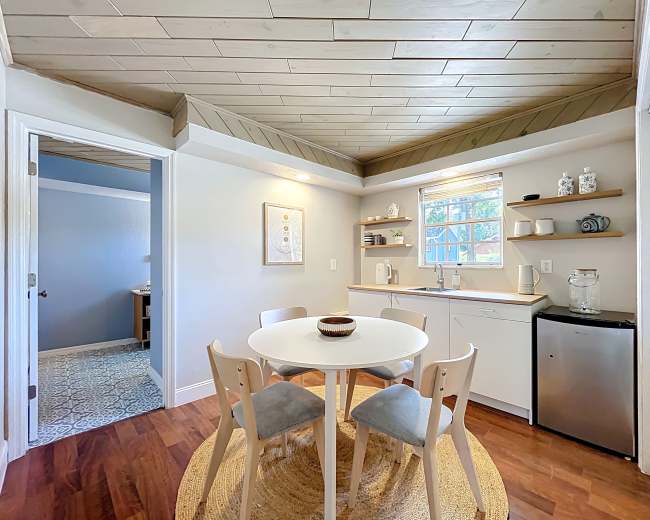 The image shows a small, modern kitchen with a round dining table, four chairs, and a window above the sink.