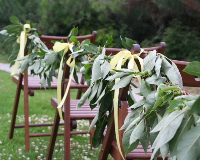 Wooden chairs are adorned with leafy garlands and yellow ribbons in a grassy area.