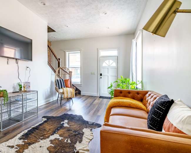 The image shows a modern living room with a brown leather sofa, a television mounted on the wall, and a plant next to a staircase.
