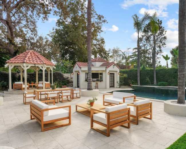 The image shows a spacious outdoor area featuring a swimming pool, a gazebo, and seating arrangements with wooden furniture surrounded by palm trees.