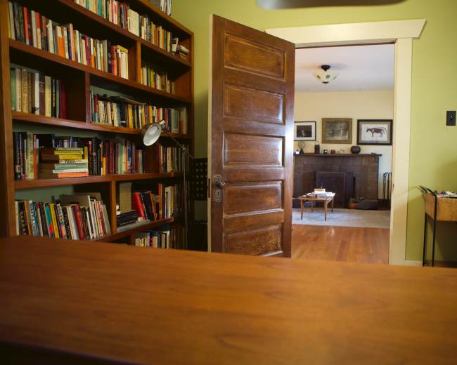 A wooden door partially opens to reveal a living room with a fireplace, framed artwork, and a coffee table, while a bookshelf filled with books is visible in the foreground.