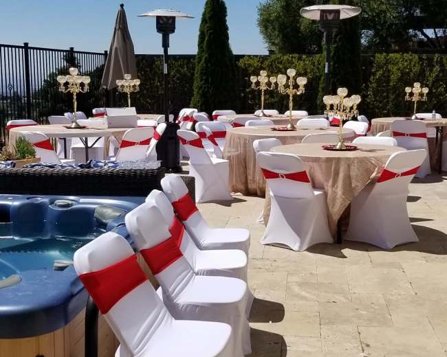 The scene features an outdoor event space with tables set for dining, each adorned with centerpieces, chairs draped in white covers with red ribbons, and a hot tub nearby.