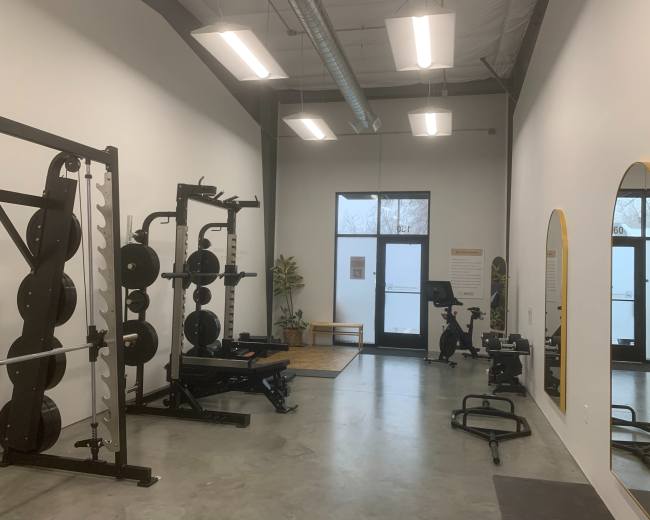 The image shows a modern gym interior featuring a weight rack, bench, exercise bike, and large mirrors, with large windows allowing natural light.