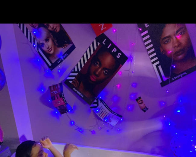 A girl relaxes in a bathtub filled with balloons and plastic balls, surrounded by colorful lights and framed images on the wall.