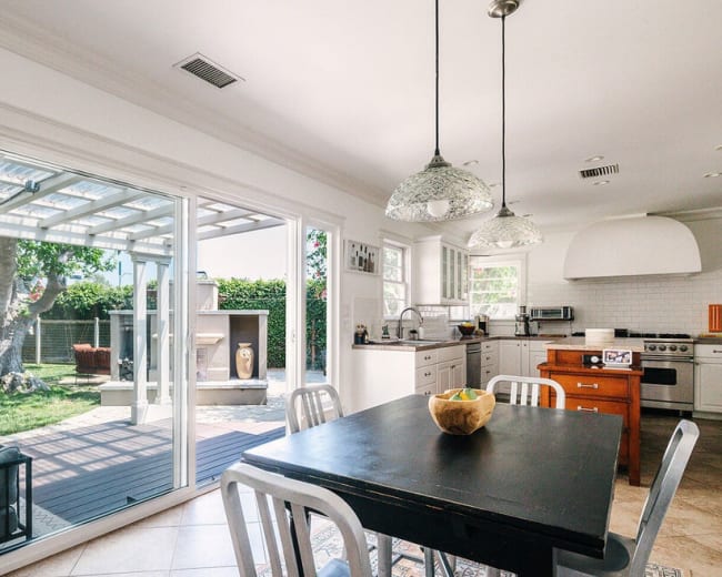 A modern kitchen features a table with black chairs, white cabinets, and sliding glass doors leading to a patio area with greenery.