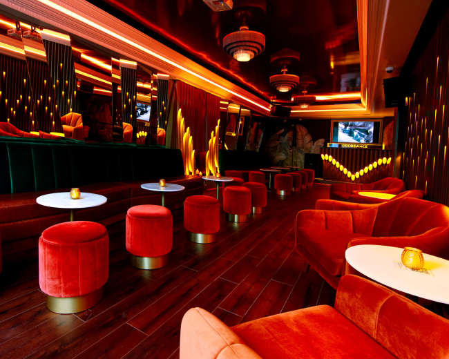 The image shows a stylish lounge with vibrant orange furnishings, plush seating arrangements, and modern lighting.