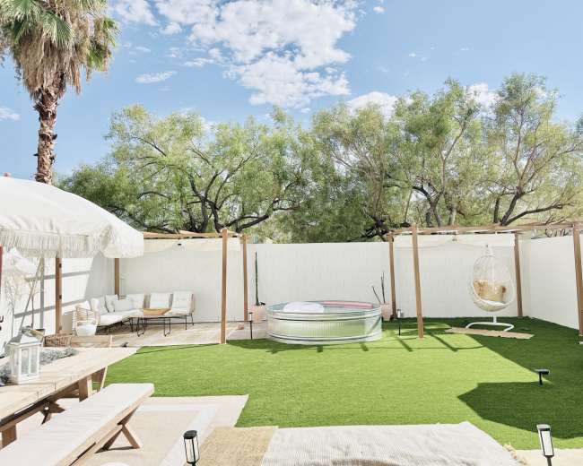 The image shows a backyard with artificial grass, a metal pool, a hanging chair, and a seating area under a shaded structure.