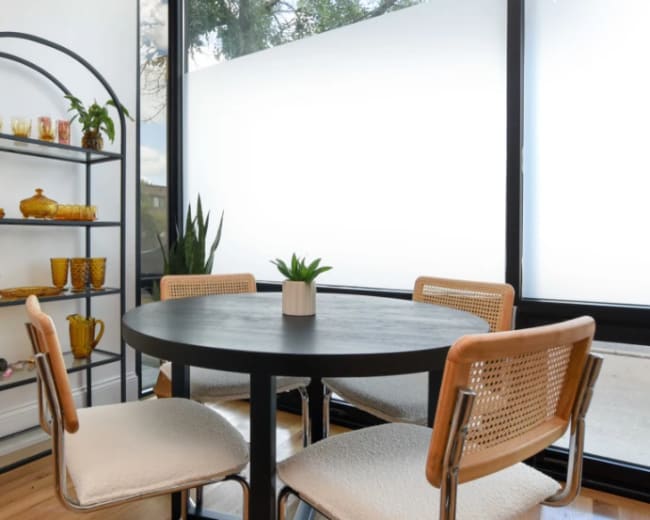A black round table is surrounded by four chairs in a well-lit room featuring a tall shelving unit with glassware and a potted plant.