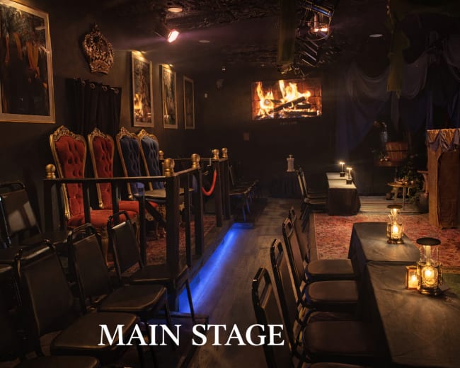 The image depicts a dimly lit theater interior featuring a main stage with red and gold throne-like chairs, a fireplace above the stage, and tables with lanterns for guests.