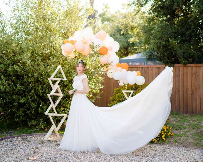A bride in a white wedding dress stands next to a balloon arch and decorative structure in a garden.