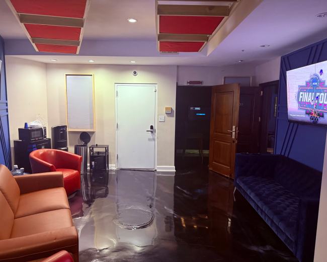 The image shows a modern lounge area with an orange leather sofa, a blue velvet couch, a red armchair, and two large screens mounted on the walls.