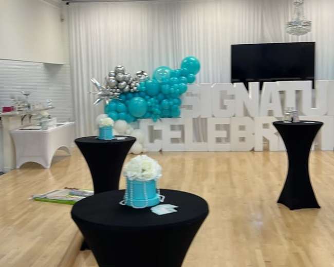 A spacious event room features black cocktail tables, a large balloon display, and a backdrop with the words "SIGNATURE CELEBRATION."