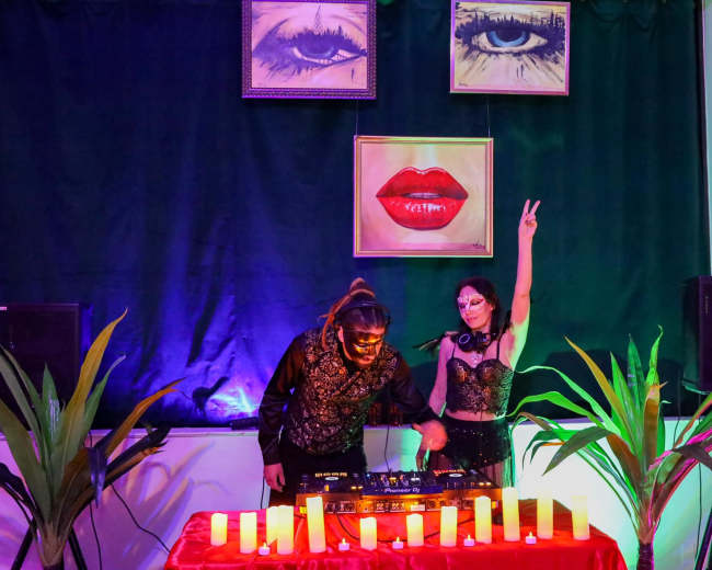 A DJ duo performs on stage in front of decorative artwork, with plants and candle lights arranged on the table.