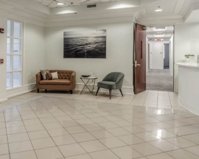 The image shows a modern reception area featuring a brown leather couch, a green chair, and a white reception desk with gray flooring and a large photograph of the ocean on the wall.