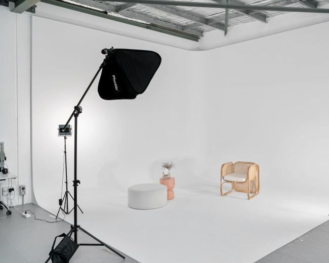 A photography studio features a simple setup with a chair, a round stool, and a potted plant against a white backdrop.