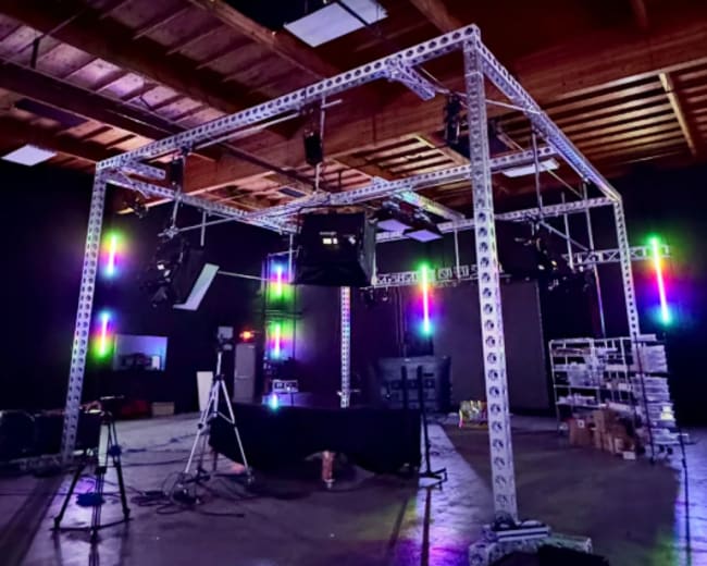 The image depicts a well-lit studio setup featuring a metal frame structure with multiple lights, cameras, and equipment arranged around a central table.