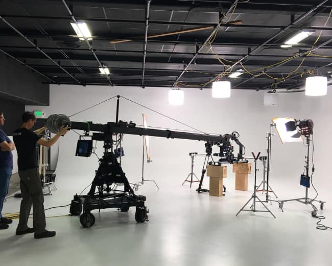 A film crew operates a camera crane in a well-lit studio set with various lighting equipment and props.