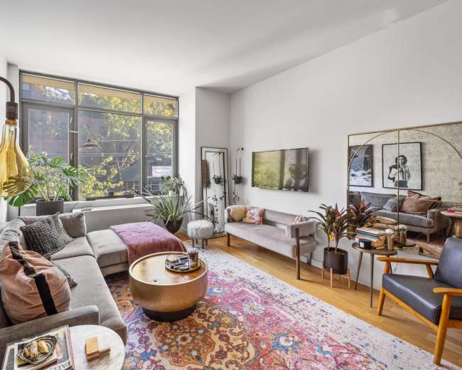 A modern living room features a gray sectional sofa, a large window with plants outside, a round coffee table, and a decorative rug.