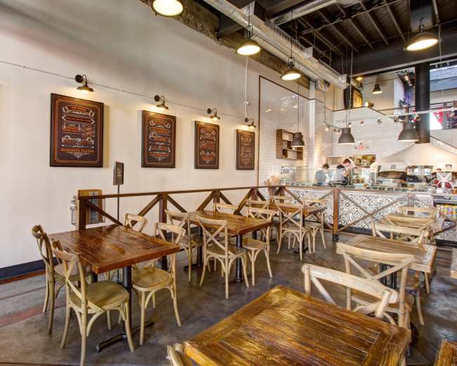 A modern café with wooden tables and chairs, exposed industrial lighting, and a menu display on the wall.