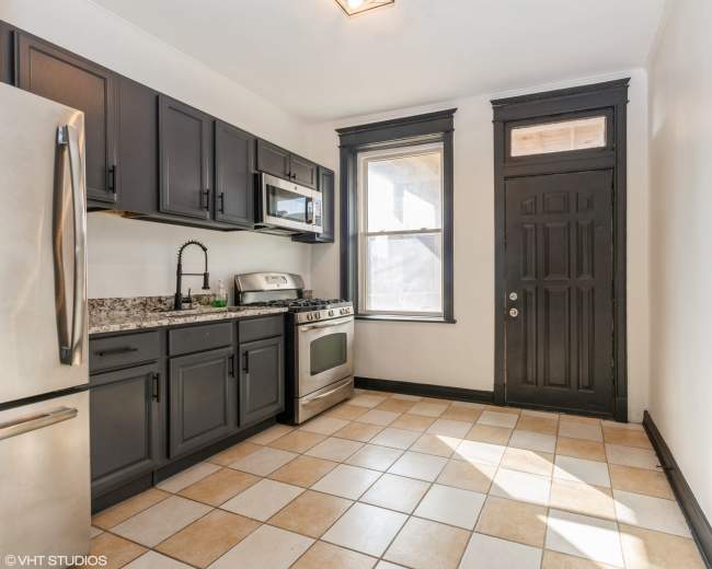 The image shows a kitchen with dark cabinets, a stainless steel refrigerator, a stove, and a tiled floor.