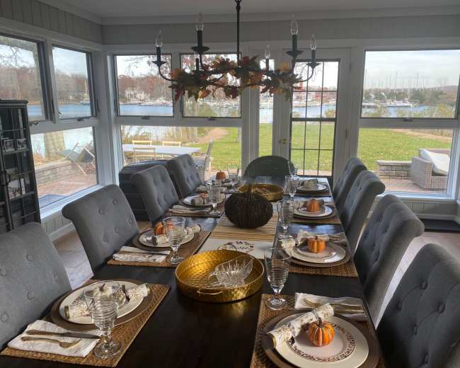 A dining room with a long wooden table set for a meal, featuring gray upholstered chairs, decorative pumpkins, and a view of a waterfront outside.