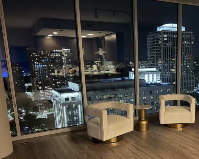 The image shows two white chairs beside a small gold table in a modern interior, with large windows revealing a city skyline at night.