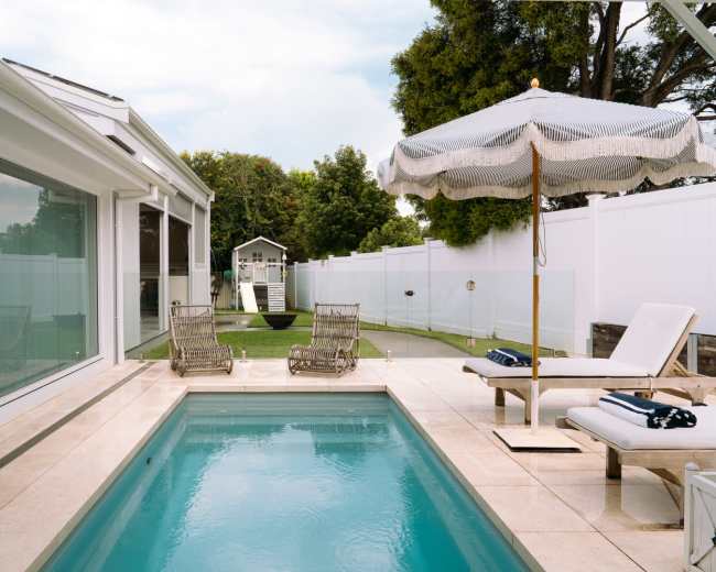 The image features a modern patio with a small swimming pool, surrounded by lounge chairs and a large umbrella, overlooking a well-maintained lawn with a playhouse in the background.