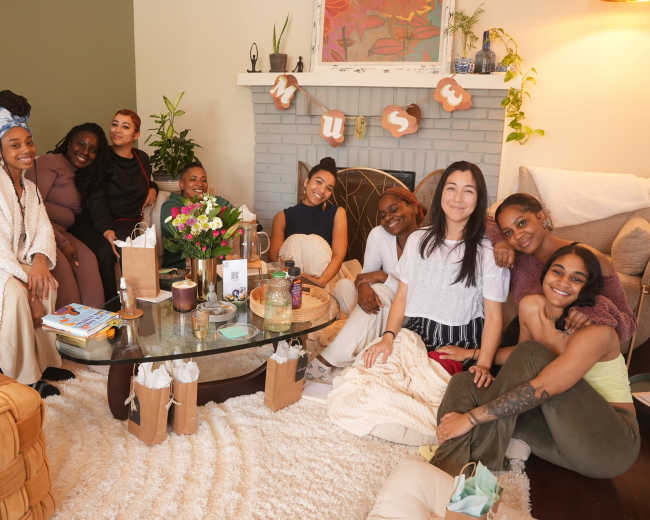 A group of nine women gathers around a coffee table in a cozy living room, smiling and enjoying each other's company.