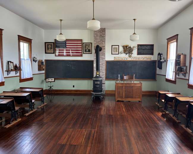The image shows a historic classroom with wooden desks, a teacher's desk at the front, chalkboards on the walls, and an American flag displayed above.