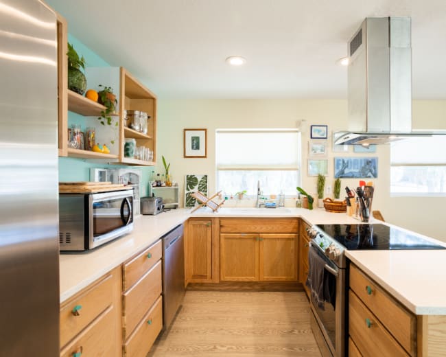 The image shows a well-lit kitchen with wooden cabinets, a central island, and various appliances and utensils visible on the countertops.
