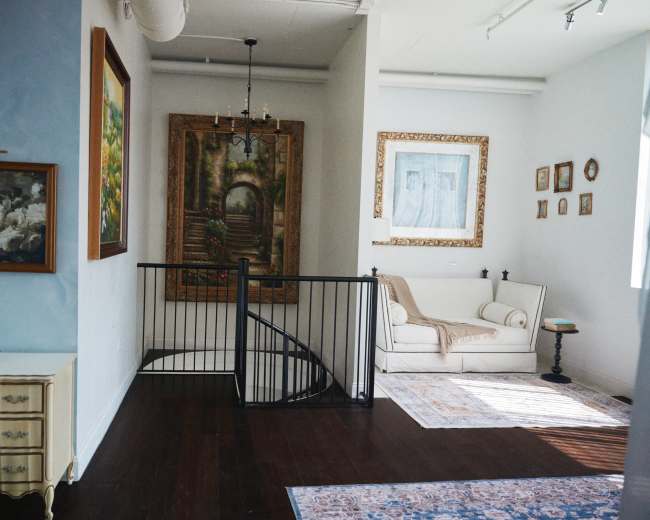 The image shows an interior space with a staircase, a white sofa, multiple framed artworks, and a small table in a well-lit room.