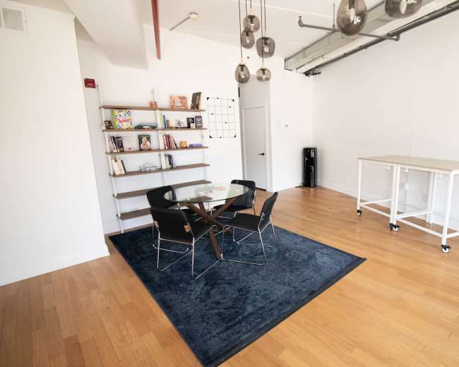 The image shows a modern, minimalistic interior featuring a wooden floor, a round table with four chairs, a bookshelf with various items, and a small white cart.
