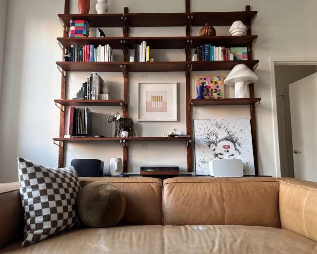 The image shows a living room scene featuring a leather couch in the foreground and a wooden bookshelf filled with various books, decorative items, and artworks in the background.