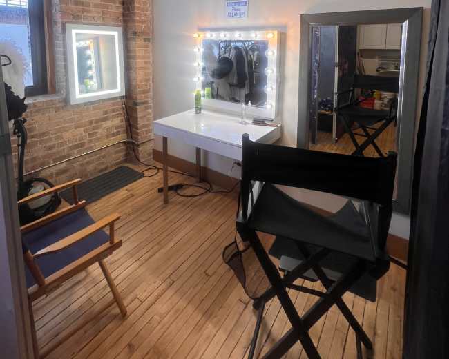 The image shows a well-lit dressing room featuring a vanity mirror with lights, a chair, and a small table, all situated against a backdrop of exposed brick walls and wooden flooring.