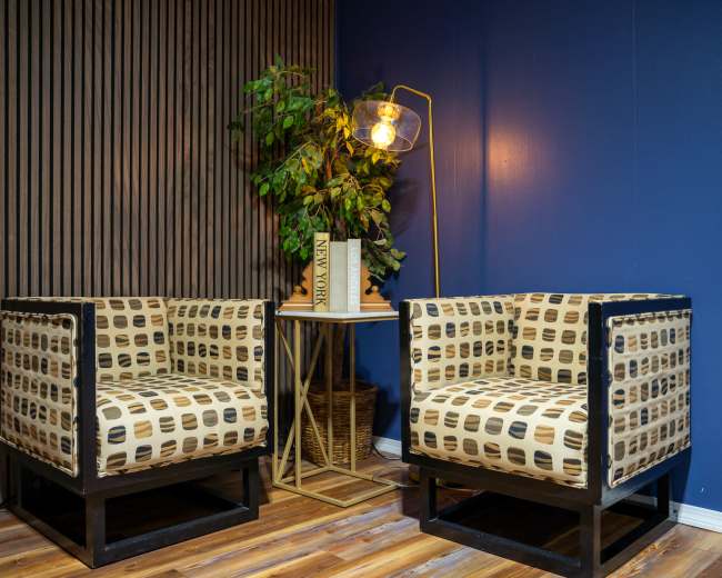 The image shows two patterned armchairs positioned next to a small table with a decorative plant and a floor lamp against a blue wall with wooden paneling.