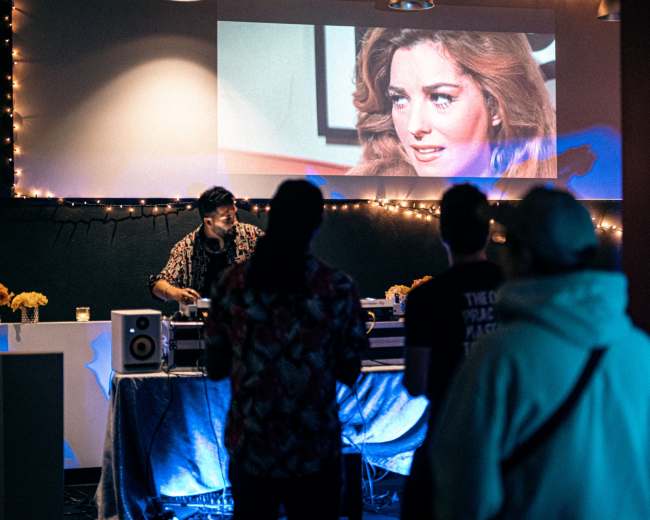 A DJ performs at a small event while an old movie scene plays on a screen behind them, with attendees watching and socializing.