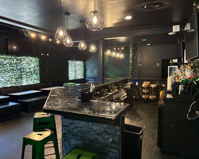 The image shows a modern bar with a marble countertop, green seating, and decorative greenery on the walls, illuminated by pendant lights.