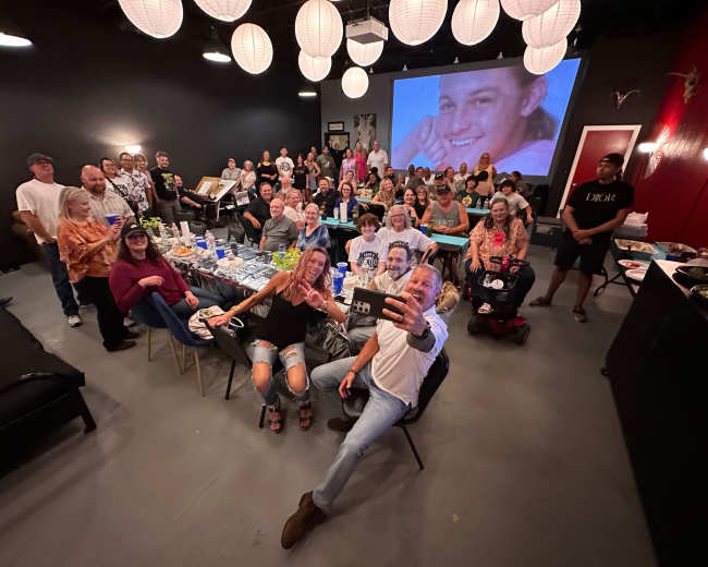 A large group of people gathers for a celebration in a dimly lit room, with a projector screen displaying a smiling face in the background.