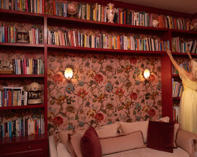 A woman in a yellow dress reaches for a book on a tall shelf lined with various books against a floral wallpaper backdrop.